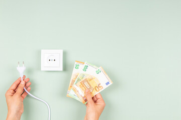 Woman hands holding electric power plug and US dollar banknotes near white electric socket on light...