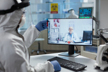 Scientist man doctor with ppe medical suit discussing medical expertise of blood infected with covid19 during online videocall telemedicine teleconference in hospital lab. Scientist virtual meeting
