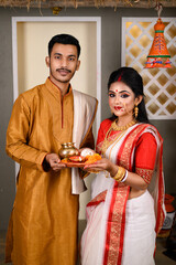 Portrait of Indian man dressed in kurta pajama with beautiful Indian woman wearing traditional Indian saree, gold jewellery and bangles, holding plate of religious offering. - obrazy, fototapety, plakaty