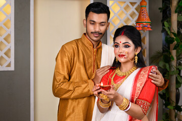 Portrait of Indian man dressed in kurta pajama with beautiful Indian woman wearing traditional Indian saree, gold jewellery and bangles, holding diya in hands on decorative background.