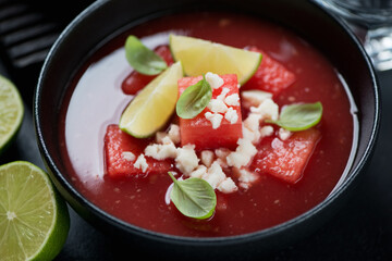 Close-up of watermelon gazpacho in a black bowl topped with bryndza and green basil leaves