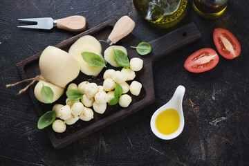 Obraz na płótnie Canvas Scamorza cheese with green basil leaves and olive oil on a black wooden serving tray, high angle view on a dark brown stone background