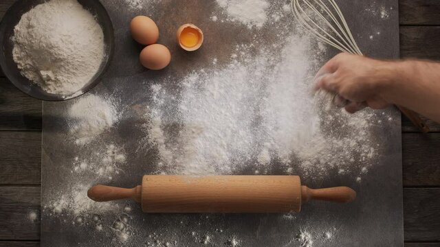 Chef's hand scatters flour on the table for rolling out the dough against the background of a rolling pin, eggs and a whisk. Home bakery concept. 