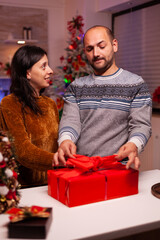 Happy couple opening xmas present gift with ribbon on it standing in x-mas decorated kitchen enjoying winter season. Cheerful joyful family celebrating winter season spending christmastime together