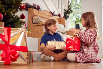 Two Excited Children Shaking And Feeling Presents Under Christmas Tree At Home