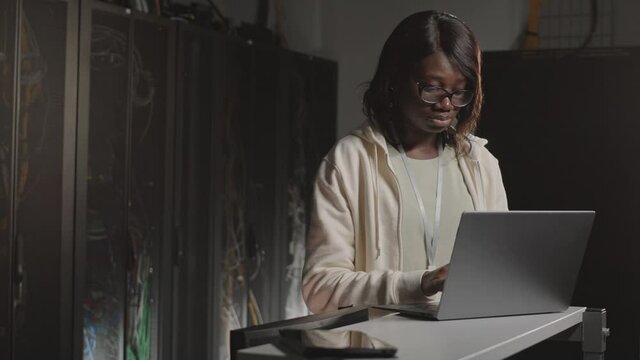 Waist-up of intelligent African American female programmer wearing eyeglasses, using laptop computer, typing in dark server room