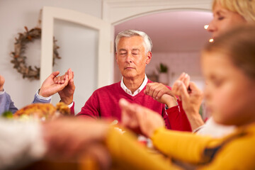 Multi Generation Family Hold Hands Around Table At Home Saying Grace Before Eating Christmas Meal