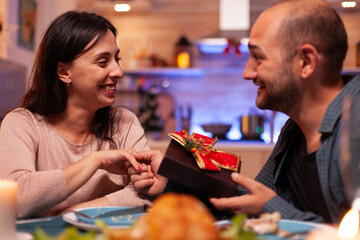 Happy couple spending christmas winter holiday surprising with xmas present with ribbon on it sitting at table in x-mas decorated kitchen. Joyful family celebrating christmastime together