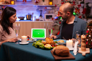 Happy family sitting at dining table in x-mas decorated kitchen enjoying winter season christmastime. Green screen mock up chroma key tablet computer with isolated display on table