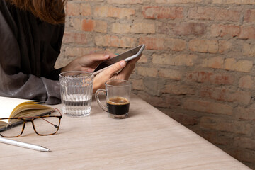 Fototapeta na wymiar Break at cafe ater work.Cups of coffee,water,glasses and notebook,pen on the wooden desk.Woman using smartphone against empty brick wall