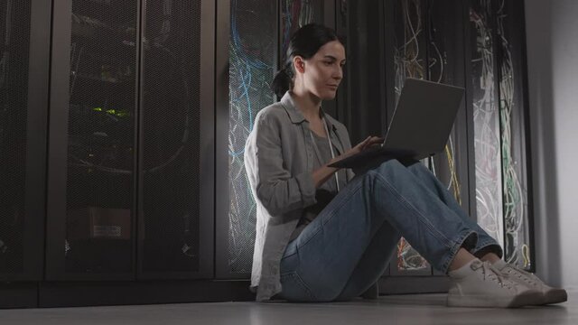 Full low angle of dark-haired Caucasian female engineer sitting on floor leaning back on racks in server room, using laptop computer, working
