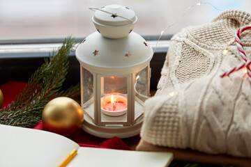 christmas, holidays and objects concept - close up of lantern with burning candle, warm wool braided sweater and sketchbook on window sill at home