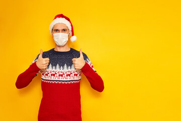 Fototapeta na wymiar man in Christmas sweater with reindeer, medical mask, New Year's hat, a Santa Claus hat, gives a thumbs up, likes, OK sign. isolated yellow background with space for text. holiday concept, gifts, sale