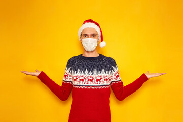 Fototapeta na wymiar excited man in Christmas sweater with reindeer, medical mask, New Year's hat, Santa Claus hat spread his hands sides, palms up, to display advertising. isolated yellow background with space for text
