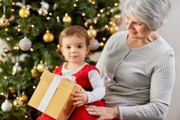 Fototapeta na wymiar christmas, holidays and family concept - happy grandmother and baby granddaughter with gift at home