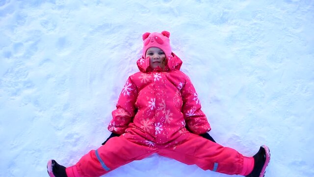 A young girl in a yellow jacket lies in the snow and makes a snow angel with his arms and legs. Child in the snow in winter. Winter outdoor games