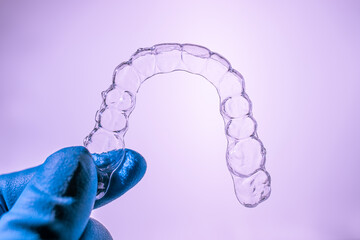 Invisible dental braces are held by a hand in a blue glove on a purple background. Plastic braces dentistry retainers to straighten teeth.