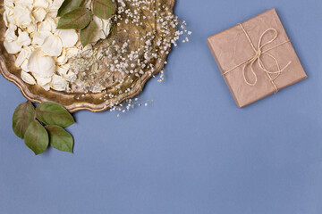 Dry petals of white roses and gypsophila on a beautiful vintage tray, a gift wrapped in craft paper on a blue background. Place for text.