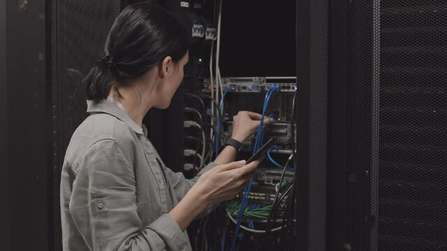 Medium close-up of dark-haired female Caucasian engineer holding smartphone, checking wire connection of server