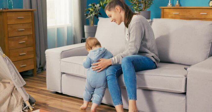 The caring mother sits with son at home, watches over him, boy stands in the living room by sofa on two legs, learns to balance, learns about the world, looks at toys, the woman belays the child