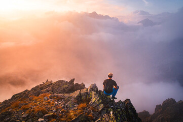 Hiker in black sit alone on the rock summit. Wonderful daybreak in mountains, rear view of backpacker sit on edge.