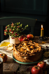 Autumn still life with fruit pie, decorated with crust apple pattern, apples,  lingonberries, candle and cup of apple tea on old wooden table and chair on background. 