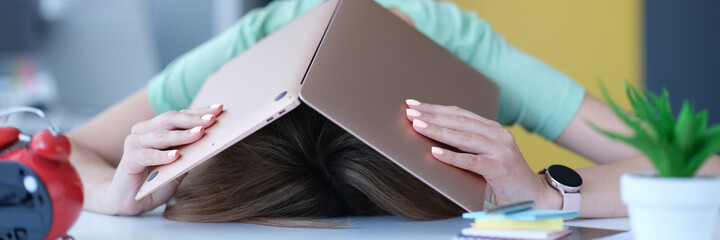 Tired in stress woman hides her head with laptop at workplace.