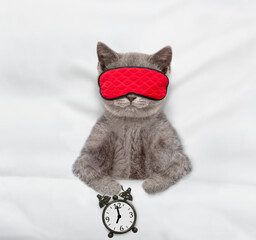 Cozy tabby kitten sleeps in sleeping mask under blanket on a bed at home with alarm clock. Top down...