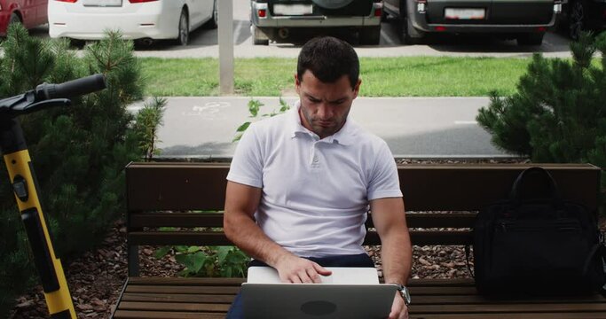 Young entrepreneur working laptop on urban bench near parking e-scooter at summer. Freelancer working remote by notebook pc on bench in city square