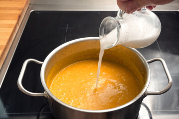 Pouring coconut milk or cream in a vegetable soup from red kuri squash or Hokkaido pumpkin in a steel pot on a black stove, cooking an autumn meal, copy space