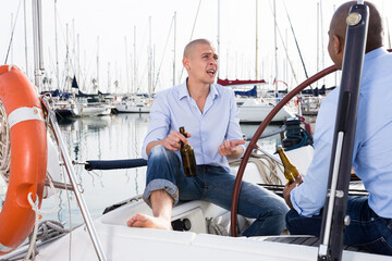 Two men in blue shirts and jeans with bare legs drinking beer and having fun on a yacht in seaport