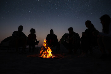Group of friends gathering around bonfire in evening. Camping season - obrazy, fototapety, plakaty