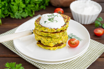 Vegetable zucchini pancakes with cheese, dill and parsley on a plate with sour cream on a dark wooden background. copy space
