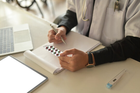 Cropped image of the doctor's hand is holding a capsule of pills and writing a prescription at the table surrounded with a white screen digital tablet and digital thermometer.
