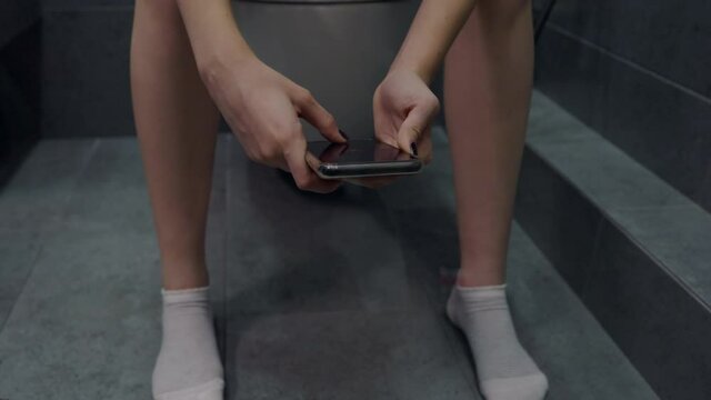 Young woman sitting on a toilet using her mobile phone