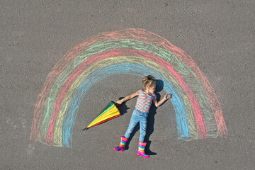 Cute little child with umbrella lying near chalk drawing of rainbow on asphalt, top view