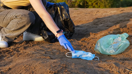 Unrecognizable woman in gloves collects trash in a black bag on the river bank, used medical masks.