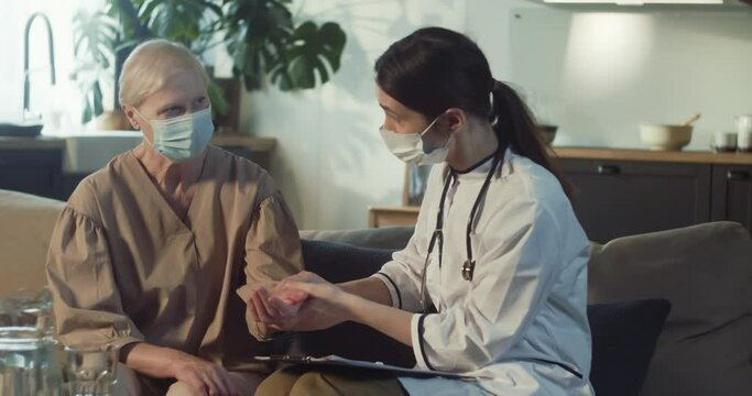 Young Female Doctor In Lab Coat Checking Pulse For 70s Senior Woman In Medical Mask At Home During House Call Visit.