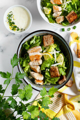 Traditional caesar salad in a white bowl on a white table with leaves and a sauce boat and parmesan. Selected focus.