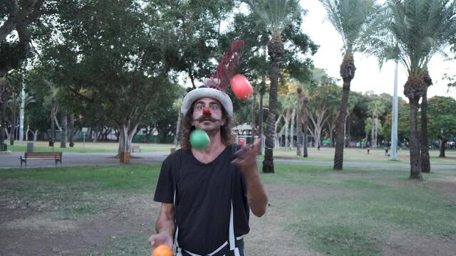 Clown with red nose Juggling Colorful Balls in the park. medium shot Steampunk dirty clown with mustache doing some circus tricks in the park. A professional circus actor juggles balls on the street.