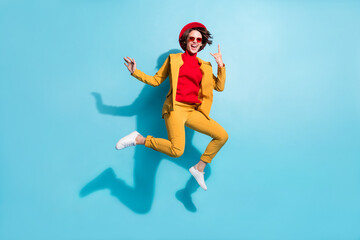 Fototapeta na wymiar Photo of pretty cool young lady wear yellow blazer red cap smiling jumping showing hard rock sign isolated blue color background