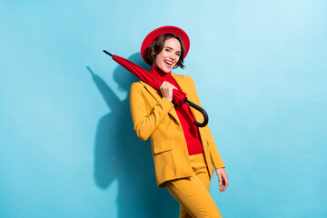 Photo of flirty adorable young lady wear yellow blazer red cap smiling holding parasol isolated blue color background © deagreez
