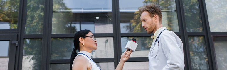 side view of asian journalist holding microphone while taking interview of doctor in white coat,...