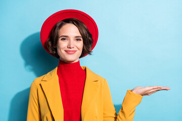 Photo of charming sweet young lady wear yellow blazer red cap smiling holding arm empty space isolated blue color background