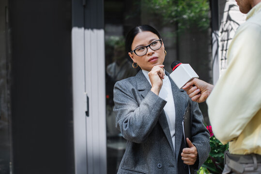 reporter with microphone near pensive asian businesswoman in glasses holding clipboard
