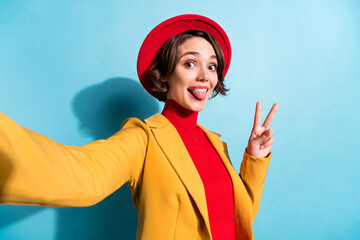 Photo of young girl happy positive smile make selfie grimace tongue-out show peace v-sign isolated over blue color background