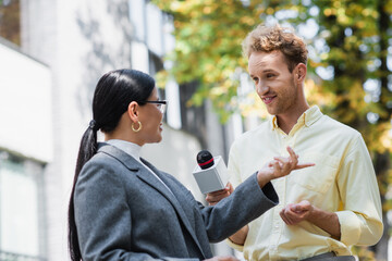 cheerful reporter holding microphone near asian businesswoman pointing with finger while giving...