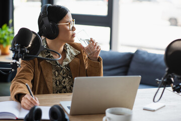 pensive asian radio host in headphones holding glass of water and pen near notebook in studio