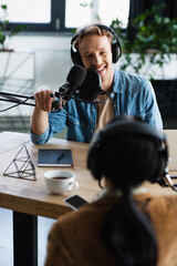 positive radio host in headphones talking in microphone and looking at blurred colleague during podcast