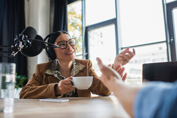 cheerful asian radio host in eyeglasses and headphones talking with colleague and holding cup during podcast